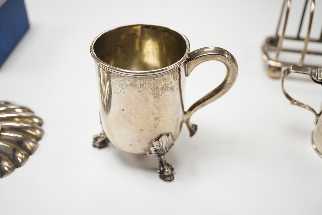 An Edwardian silver mug, on claw and hoof feet, William Hutton & Sons, London 1909, height 83mm, together with a silver butter shell, mustard pot and a silver five bar toastrack, 17oz.